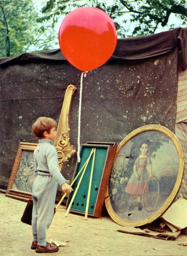 Der rote Ballon, Szenenbild: Ein kleiner, grau gekleideter Junge steht mit einem großen, roten Luftballon vor einem Gemälde. Es zeigt ein Mädchen mit einem Reifen.