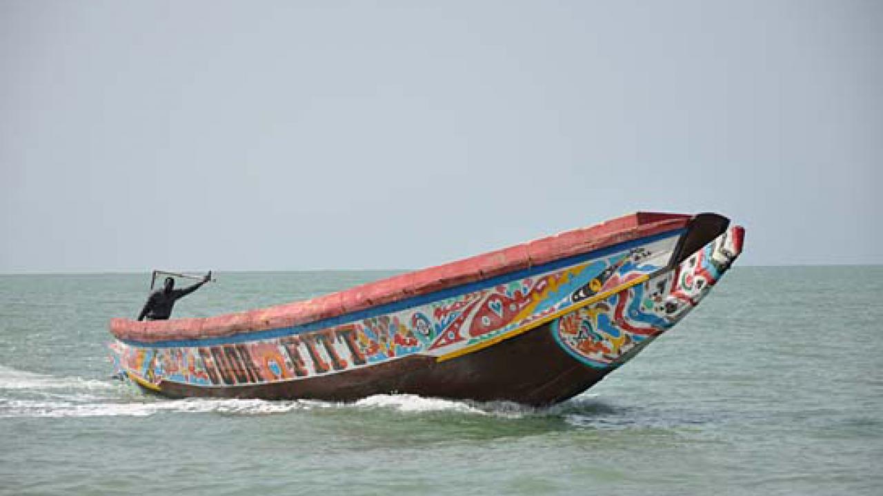 Szenenbild aus dem Film <filmtitel>Die Piroge</filmtitel>: Ein einfaches, bunt bemaltes Boot fährt im Meer. Ein Mann sitzt am hintren Ende und hebt seinen linken Arm nach oben.