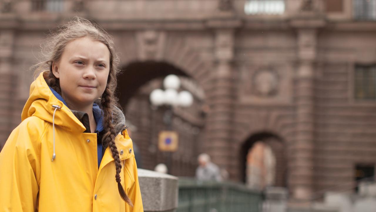 Greta Thunberg steht mit geflochteten Haar und in einem gelben Regenmantel vor einem Gebäude Stockholm. Sie guckt aufgeschlossen vor sich hin. 