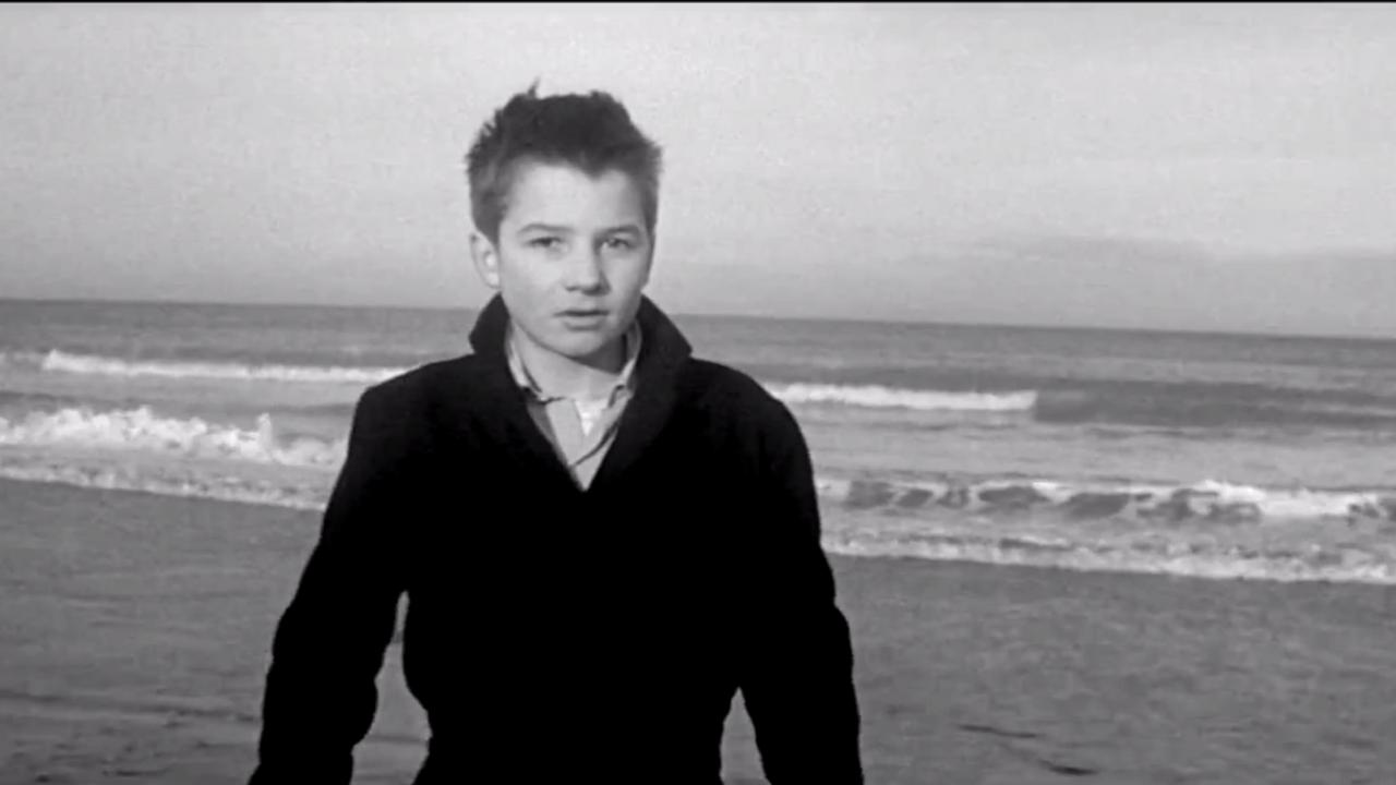 Nahaufnahme eines Junge am Strand in Schwarz-Weiß, er blickt direkt in die Kamera. Hinter ihm ist das Meer. 