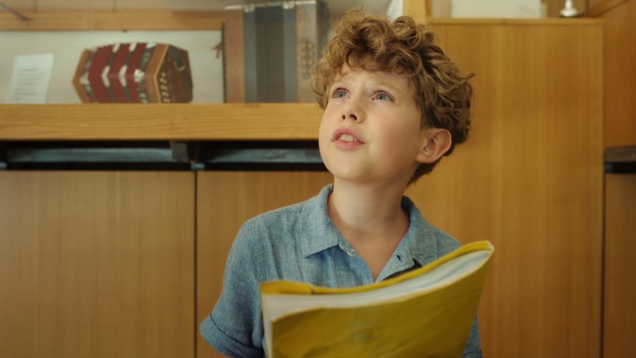 Nahaufnahme eines Jungen mit lockigen Haaren und blauen Hemd, der leicht nach oben schaut. Er hält ein gelbes Heft ind den Händen. Hinter ihm sind Holzschränke. 