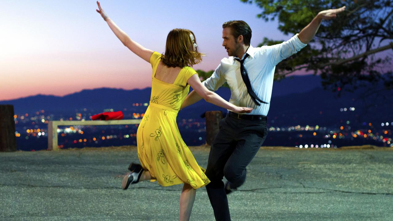 Szenenbild aus dem Musical La La Land: Eine junge Frau und ein junger Mann halten sich an den Hüften und tanzen mit erhobenen Armen. 