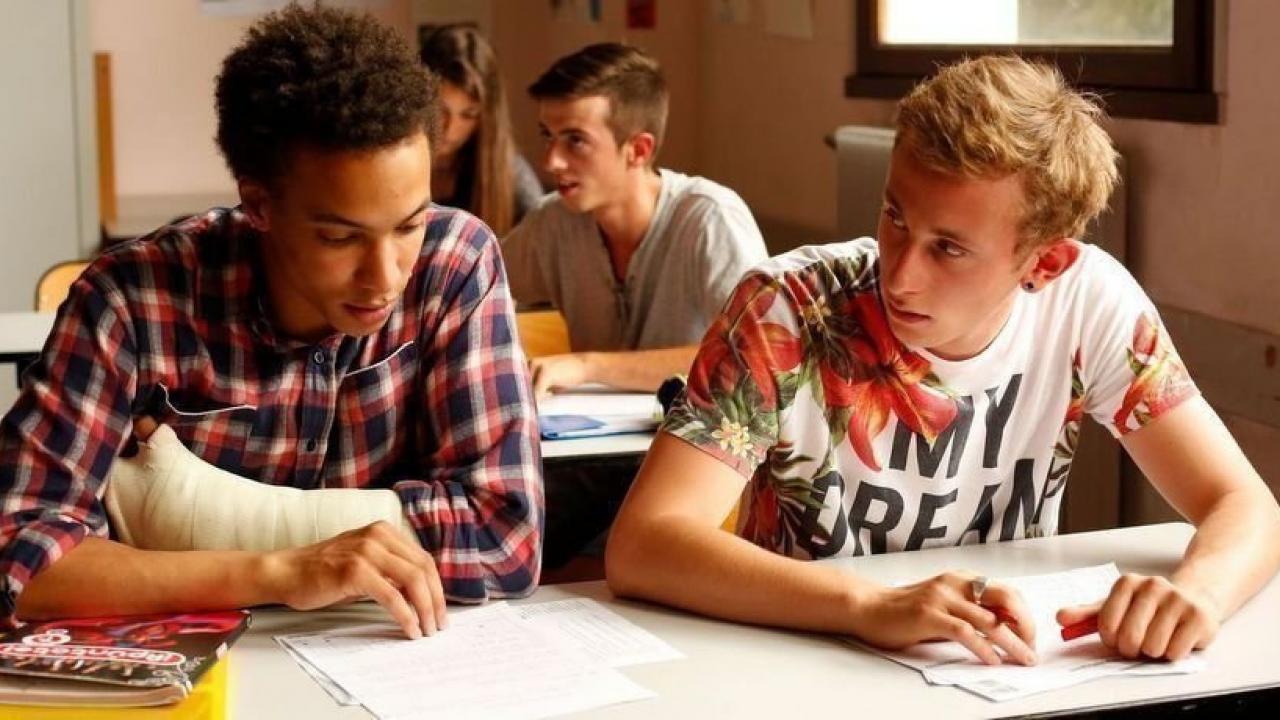 Zwei Jugendliche sitzen in der Schule am selben Tisch. Links ein junge mit dunklen Haare, karrierten Hemd und linke Hand im Gips. Er guckt nach unten auf seine Schulnotizen. Rechts ein blonder Junge mit weißen T-shirt mit Blumen. Darauf kann man "My dream" in schwarzer Schrift lesen. Er guckt den anderen Jungen an.