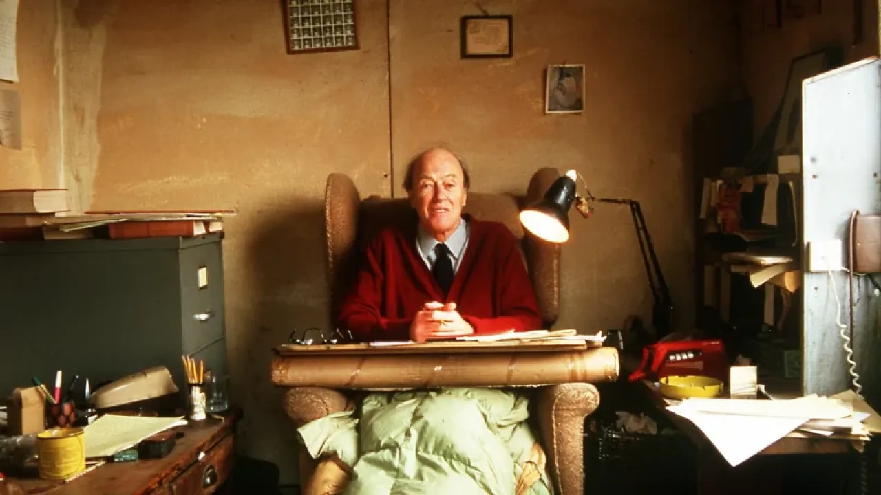 Der Autor Roald Dahl sitzt mit roten Pulli und grüner Decke in seinen Arbeitsstuhl. Rechts von ihm steht eine Lampe, Links ein Dokumentenkabinett. Mit den Händen vor ihn, schaut er lächelnd in die Kamera hinein.