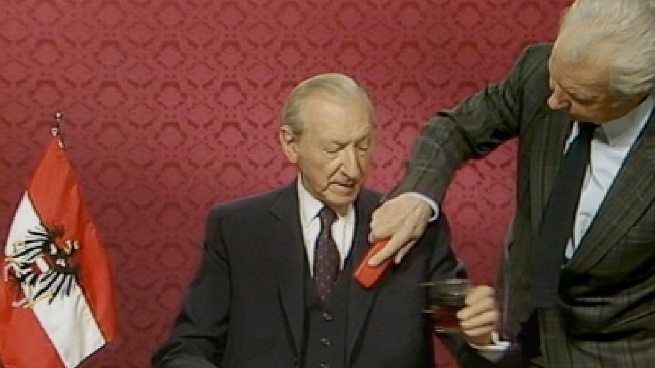 Der österreichische Politiker Kurt Waldheim kurz vor eine Rede im Fernsehen. Jemand bürstet ihm die Jacke ab. Er hält ein Glas in seiner Hand. 