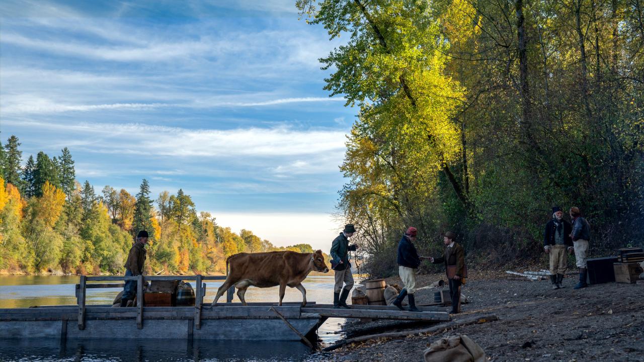 Szenenbild aus dem Film "First Cow": Totale einer Flußlandschaft. Ein Mann führt eine braune Kuh von einem Boot aus ans Land.