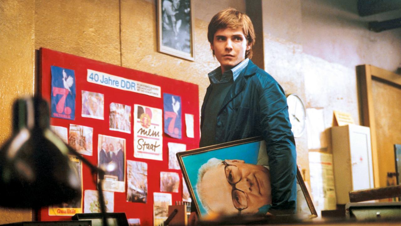 Szenenbild aus dem Film "Good Bye, Lenin!": Ein junger Mann steht vor einer Tafel mit Bilder, die die Überschrift "$0 Jahre DDR" trägt. Er hält ein Bild von Erich Honecker unterm Arm.