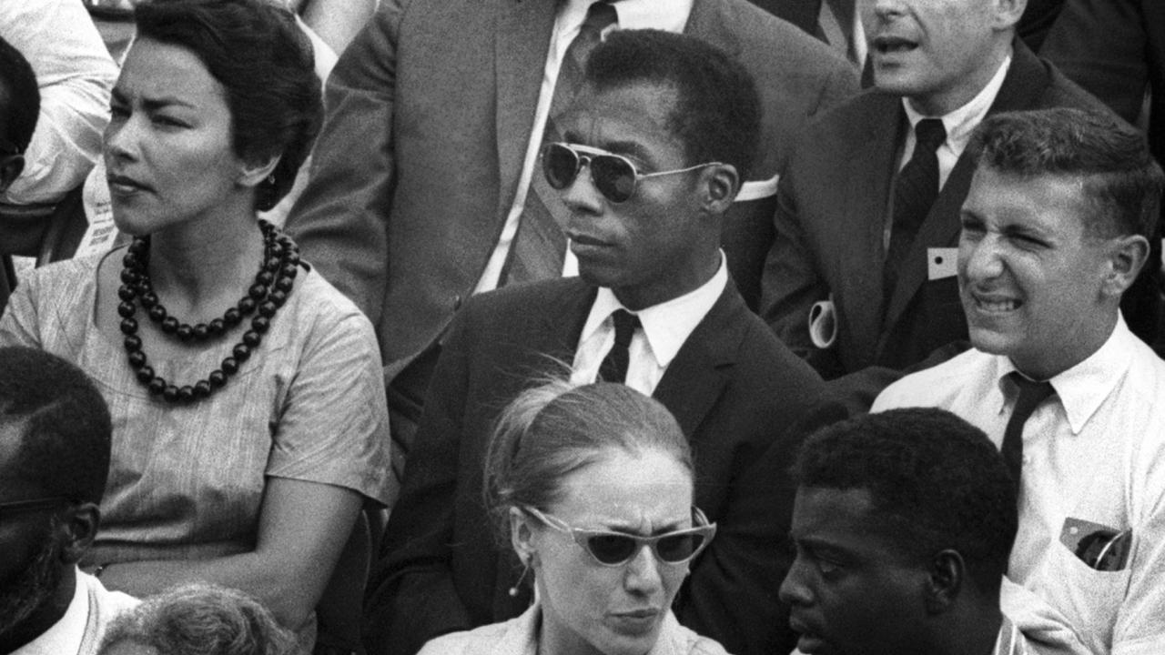 I Am Not Your Negro, Szenenbild: Halbnahe Einstellung eines Publikums. In der Mitte sitzt der afroamerikanische Schriftsteller James Baldwin mit Sonnenbrille. Das Bild stammt aus de 1960ern.