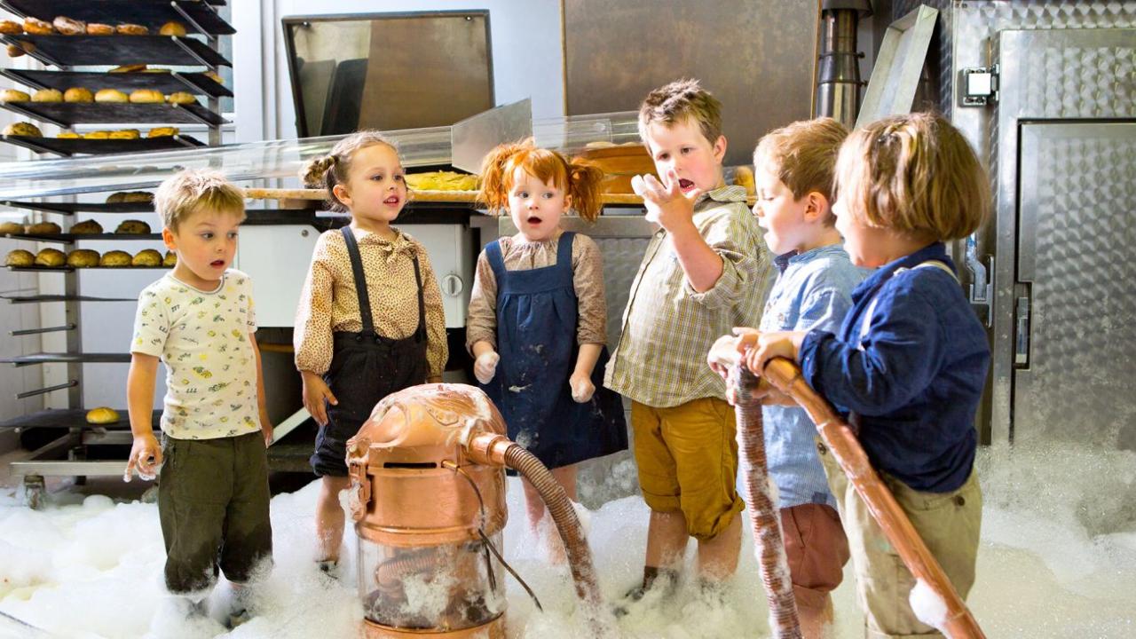 Sechs Kinder stehen in der Reihe in einer Bäckerei, vor ihnen ist ein rauchender Staubsauger