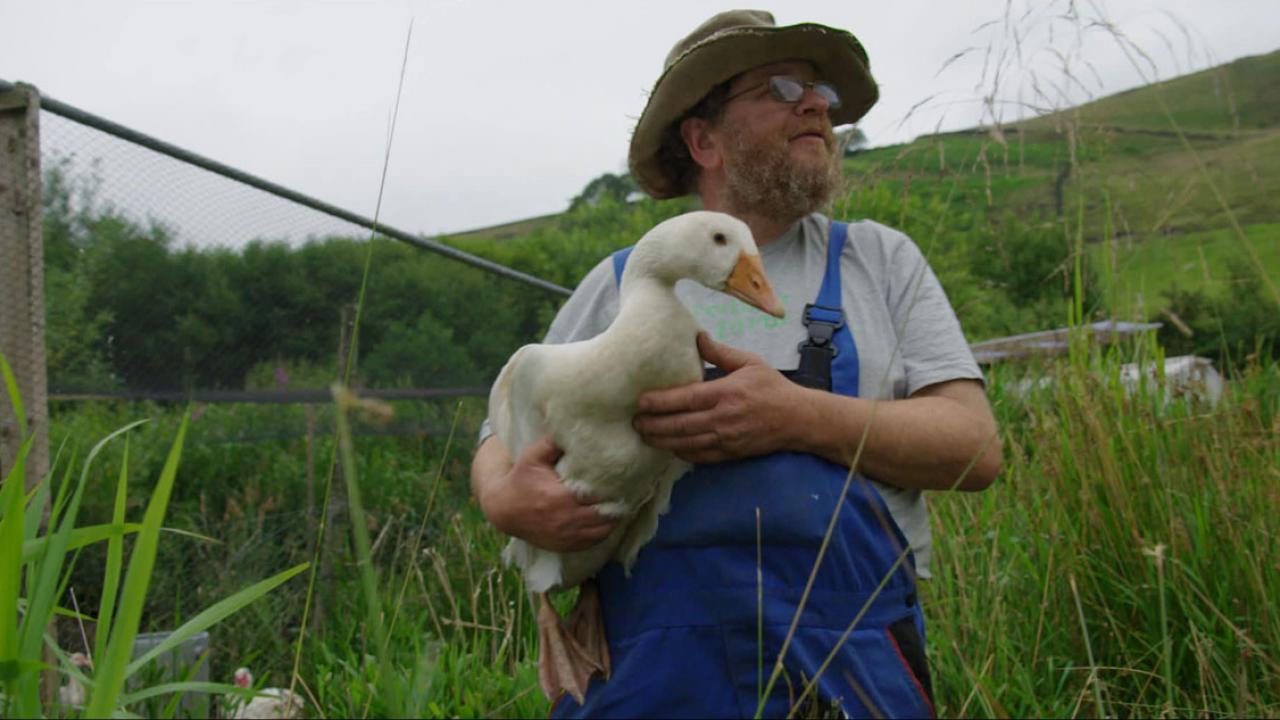 Ein Mann in blauer Latzhose, Brille und Hut, steht in im Gras und hält eine Gans im Arm. 