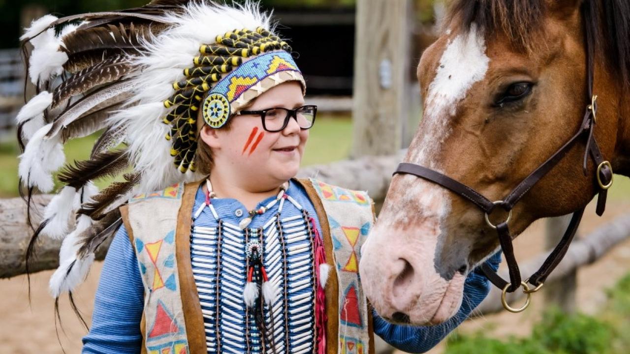 Kind im Indianerkostüm mit Federschmuck streichelt ein braunes Pferd.