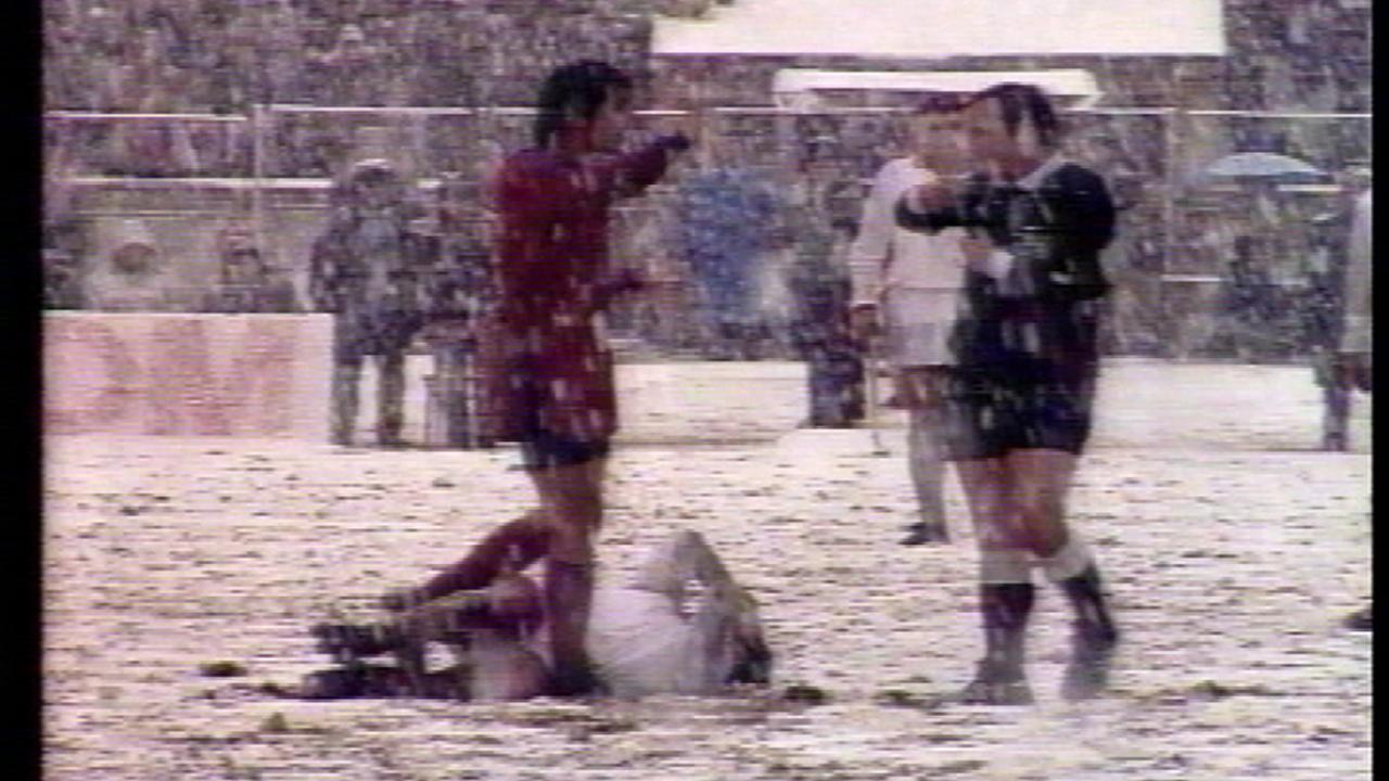 Auf einem verschneiten Fußballfeld stehen zwei Männer in Fußballkleidung und ein weiteren Mann liegt am Boden, in Schmerzen. Es schneit stark und das Bild ist schlecht. 