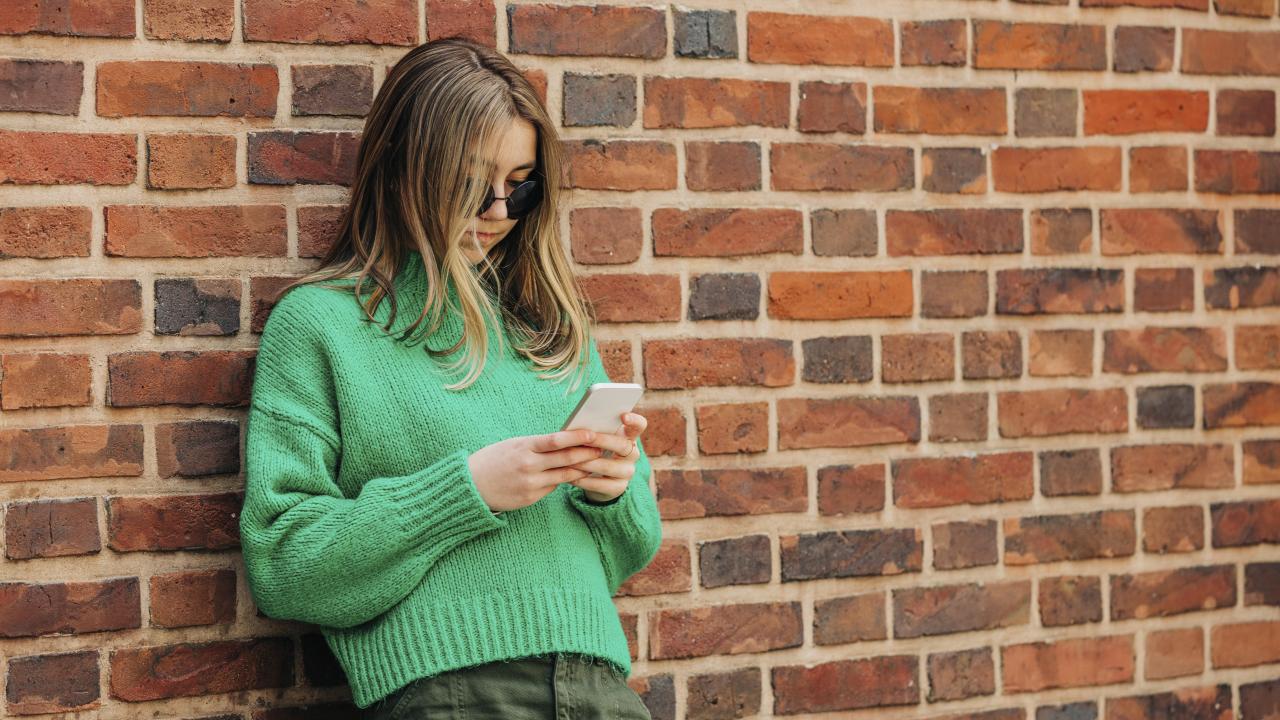 Eine Jugendliche mit Sonnenbrille lehnt sich an eine Backsteinmauer und schaut auf ihr Smartphone