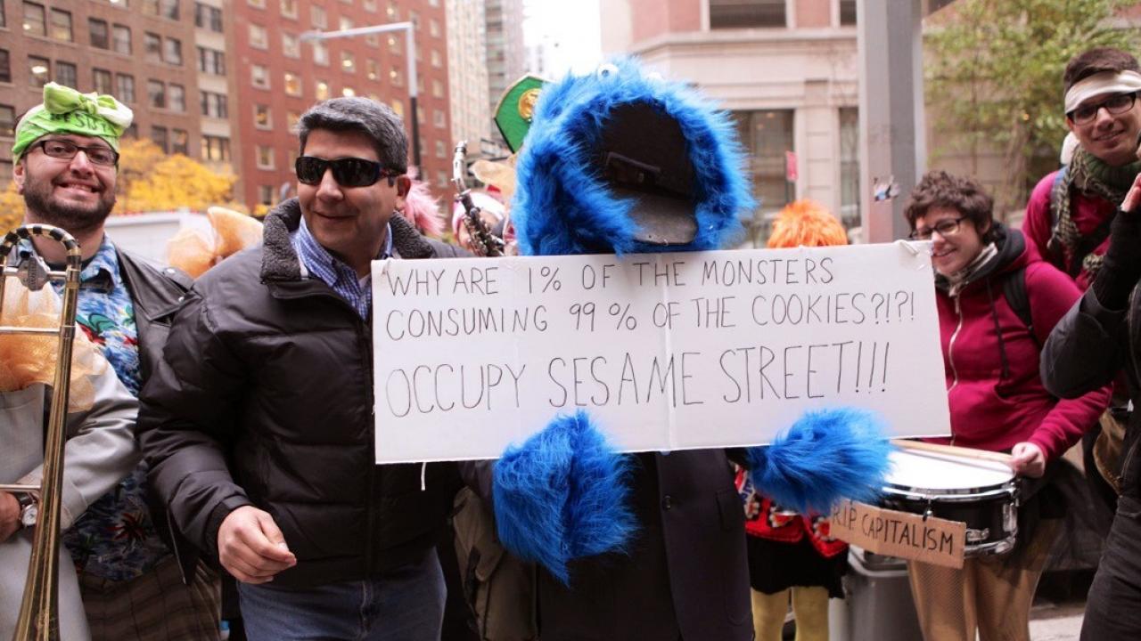 Gruppe von Menschen, darunter eine Person im Krümelmonster-Kostüm, hält ein Schild mit der Aufschrift "Why are 1% of the monsters consuming 99% of the cookies?!?! Occupy Sesame Street"