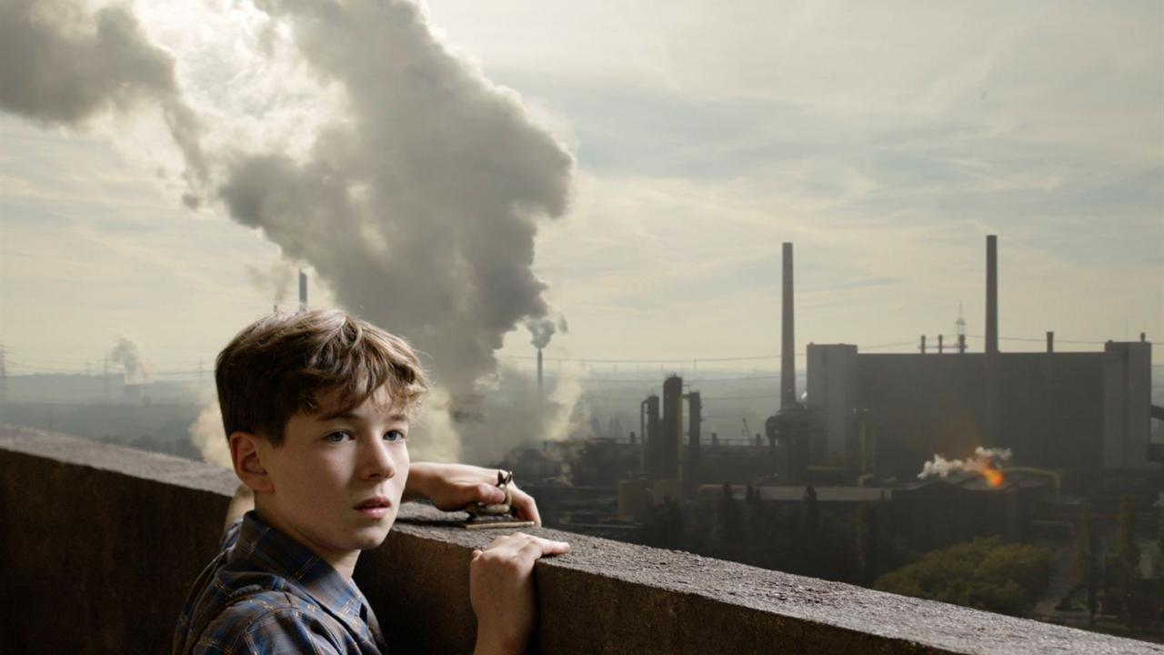 Ein Junge steht an einer Mauer und schaut besorgt auf eine rauchende Fabriklandschaft. Industrielle Gebäude und Schornsteine im Hintergrund.