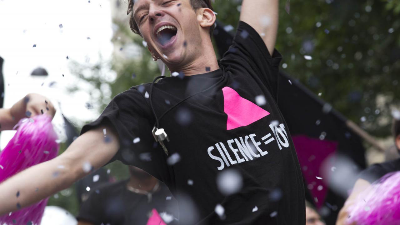 Ein jubelnder Mann in einem T-Shirt mit der Aufschrift "Silence = Death" tanzt inmitten von Konfetti und Feiernden.