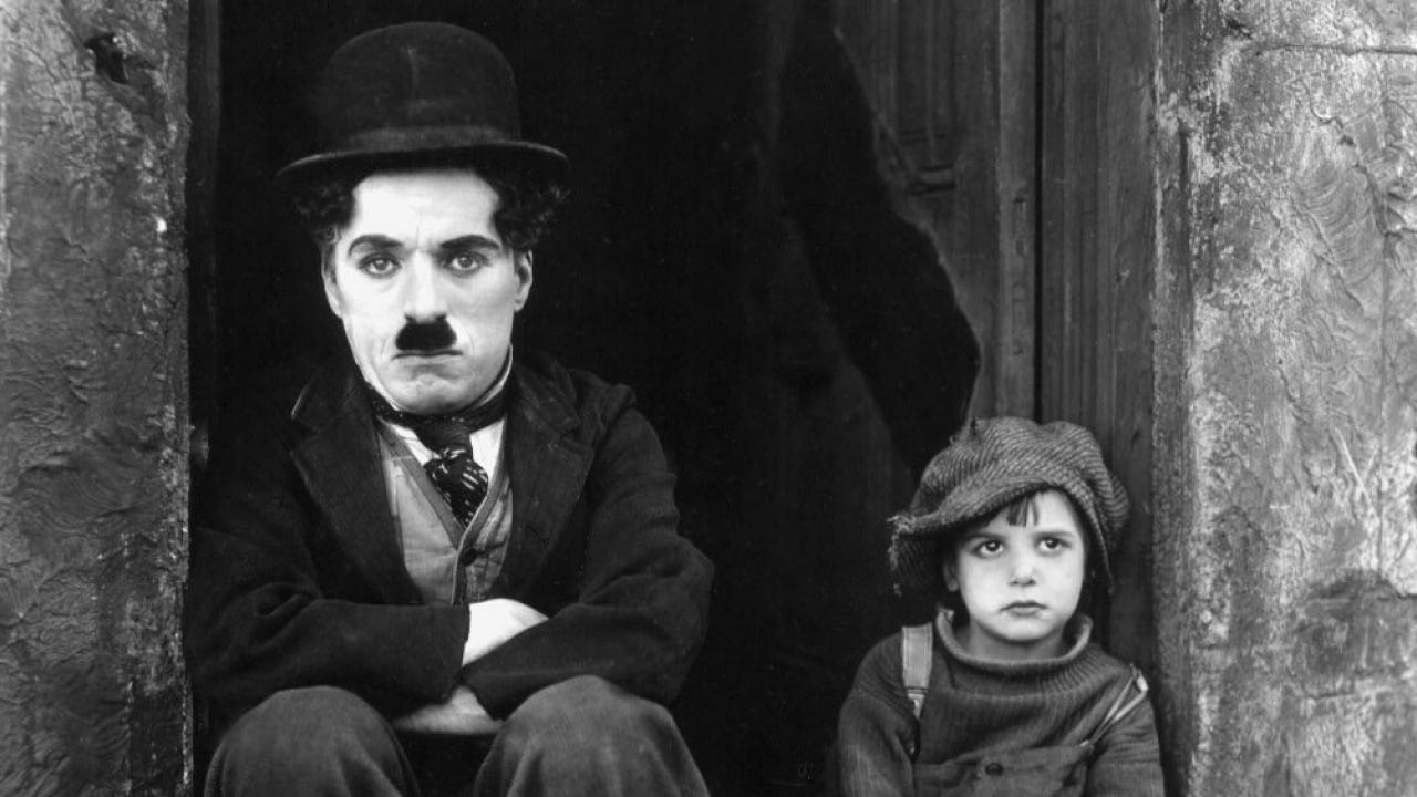 Schwarz-Weiß-Foto von Charlie Chaplin und einem Jungen in zerlumpter Kleidung, beide sitzen vor einer Tür und schauen ernst.