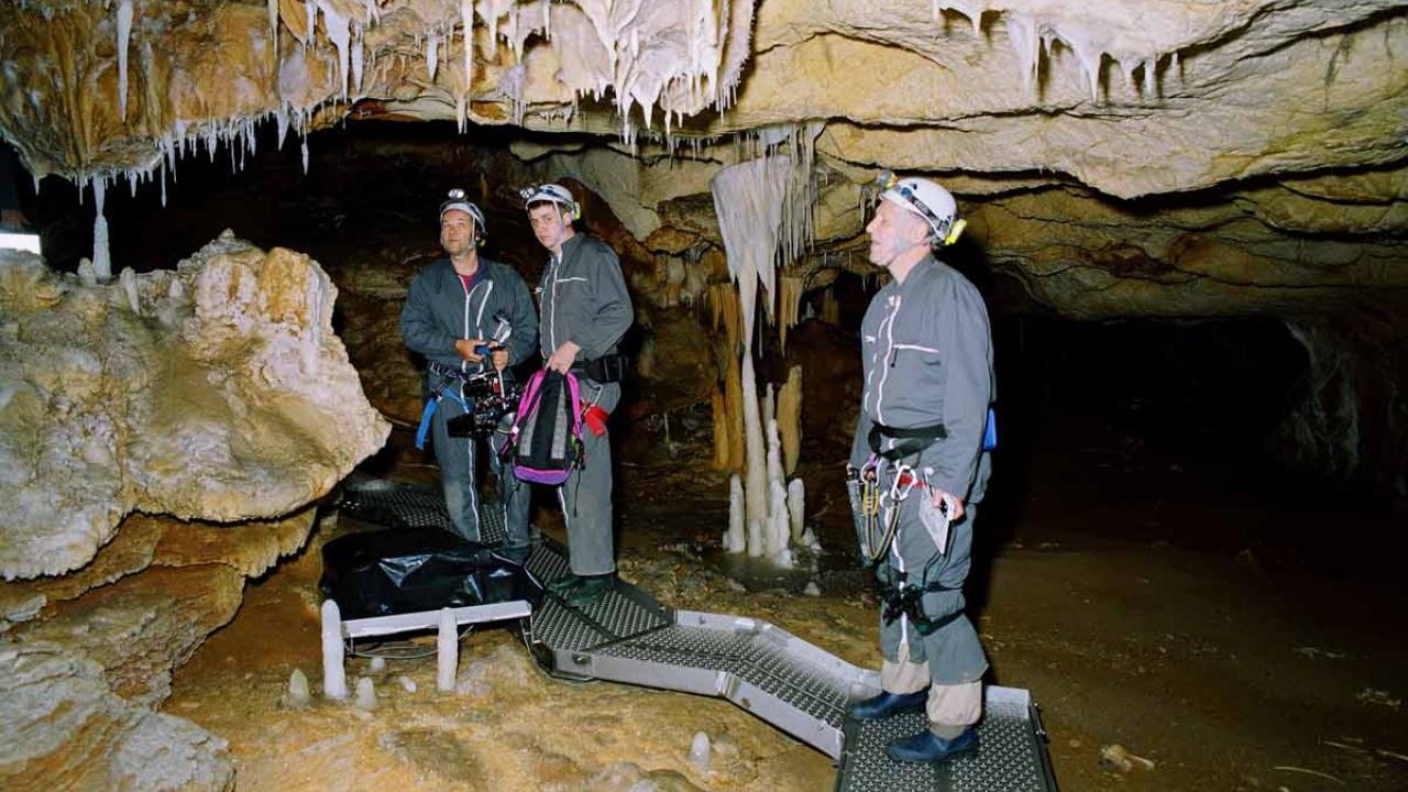 Drei Höhlenforscher stehen in einer Tropfsteinhöhle, umgeben von Stalaktiten und Stalagmiten.