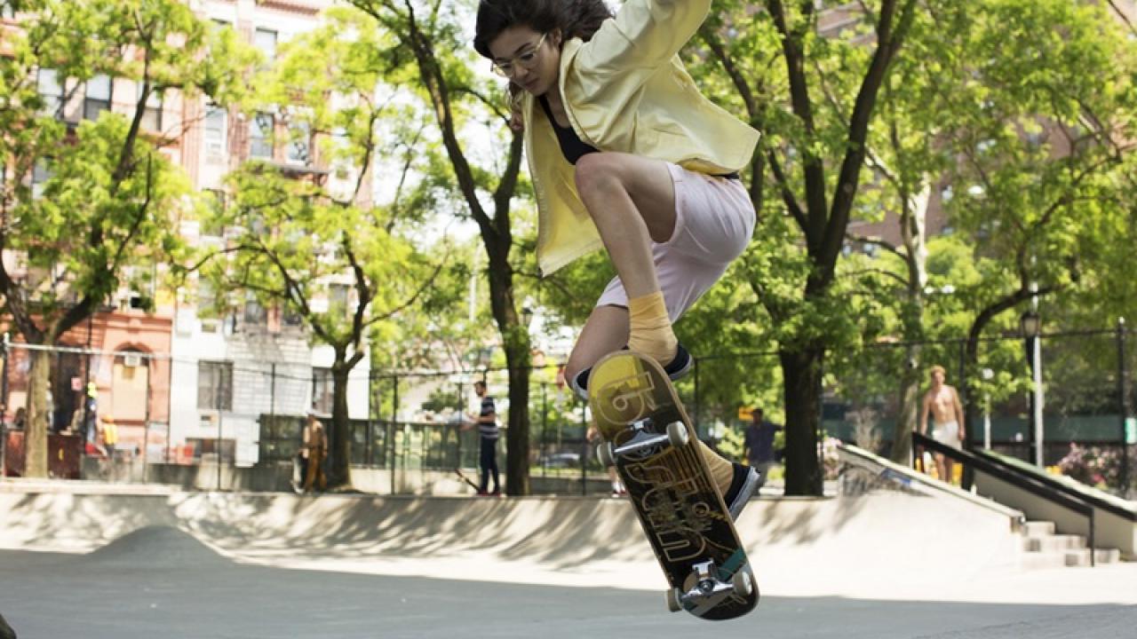 Junges Mädchen in gelber Jacke und rosa Shorts macht einen Skateboard-Trick im Skatepark.