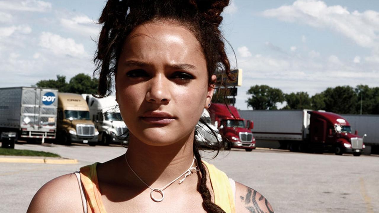 Frau mit Dreadlocks und Tanktop steht auf einem Parkplatz mit LKWs im Hintergrund. Sonnenlicht und blauer Himmel.