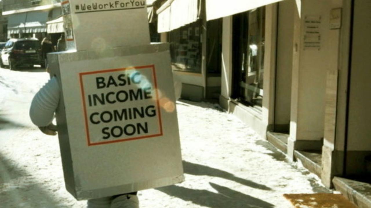 Person im Kostüm aus Pappkartons mit der Aufschrift "Basic Income Coming Soon" läuft auf schneebedeckter Straße.