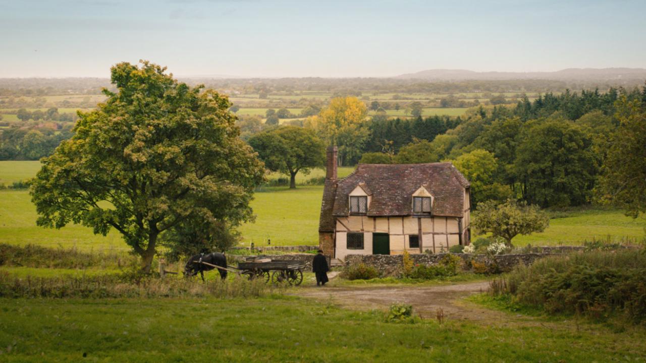 Totale eines kleinen Hauses mit Reetdach in einer grünen, hügeligen Landschaft. Links neben dem Haus steht ein belaubter Baum, ein Weg führt zu Haustür.