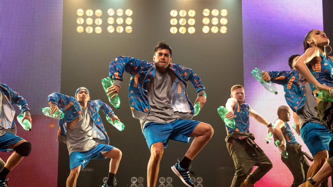 Gruppe von Tänzern in blauen Outfits performt energetische Choreografie auf Bühne mit Flaschen, bunte Lichter im Hintergrund.