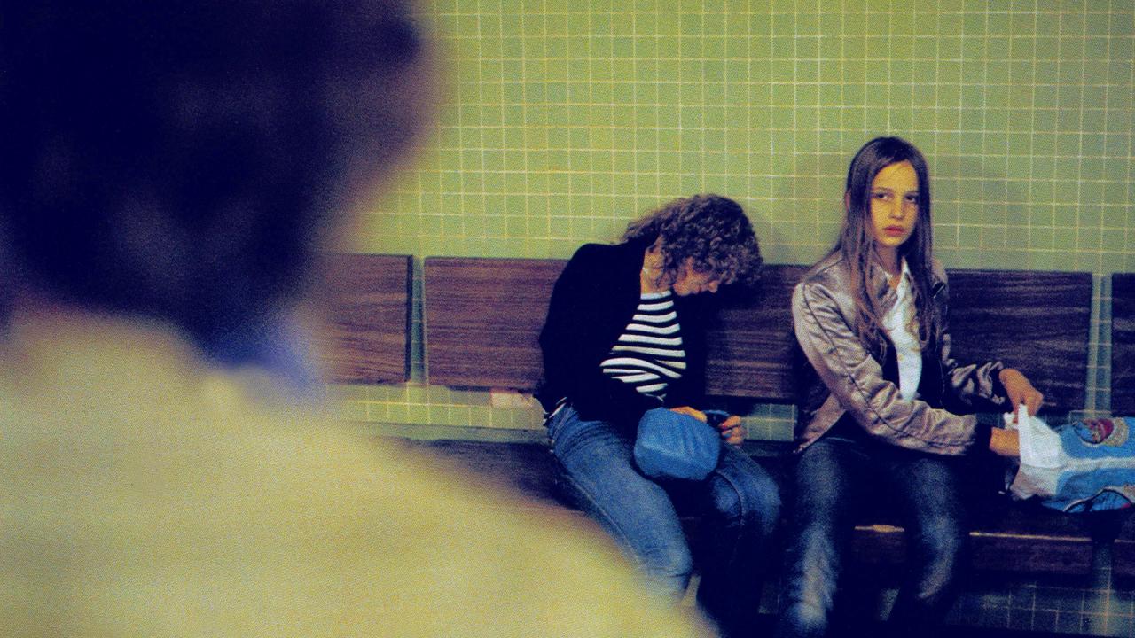 Szenenbild aus dem Film CHRISTIANE F. – WIR KINDER VOM BAHNHOF ZOO: Zwei junge Mädchen sitzen auf einer Bank in einem U-Bahnhof. Das eine Mädchen ist in sich zusammengesackt. Im Vordergrund ist angeschnitten die Silhouette eines Mannes zu erkennen, der in Richtung der beiden Teenagerinnen schaut.