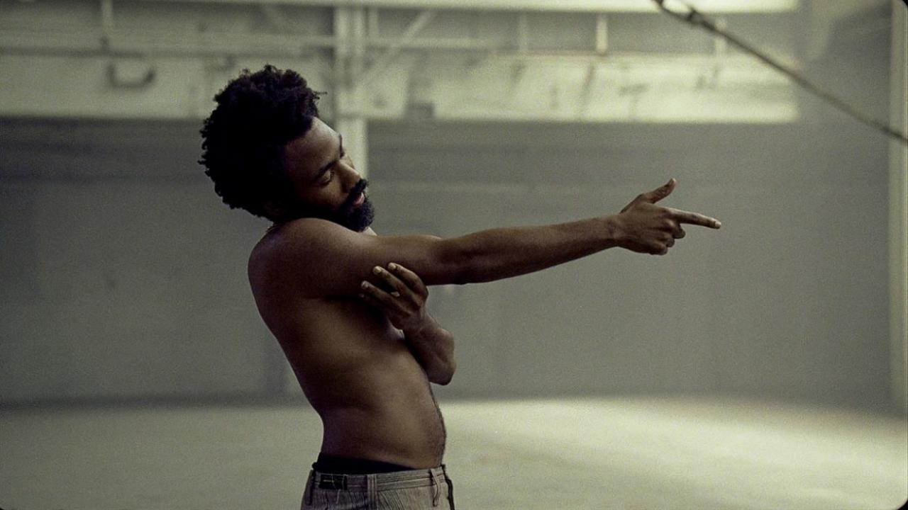 This is America, Szenenbild: Nahaufnahme des afroamerikanischen Musikers Childish Gambino. Er steht mit nacktem Oberkörper in einer Halle und zielt mit dem Finger nach rechts.