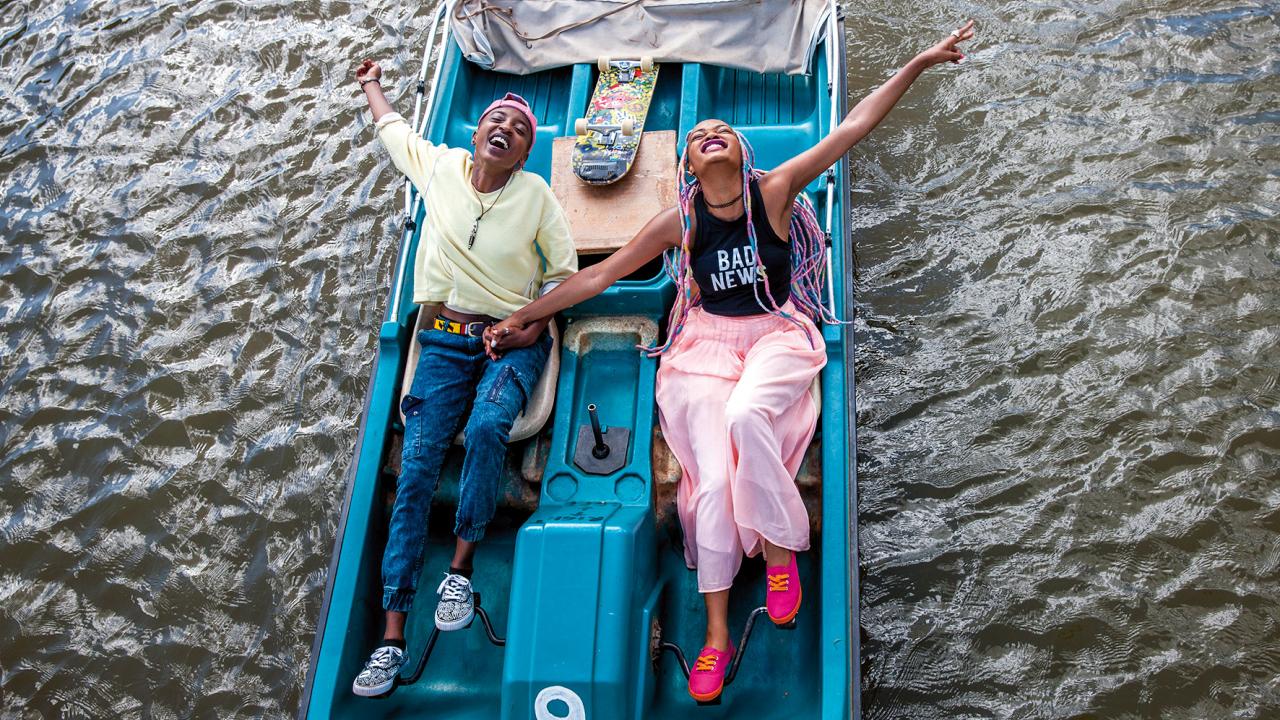 Szenenbild aus dem Film RAFIKI: Zwei junge Schwarzen Frauen setzen un einem Trettboot und lachen. 