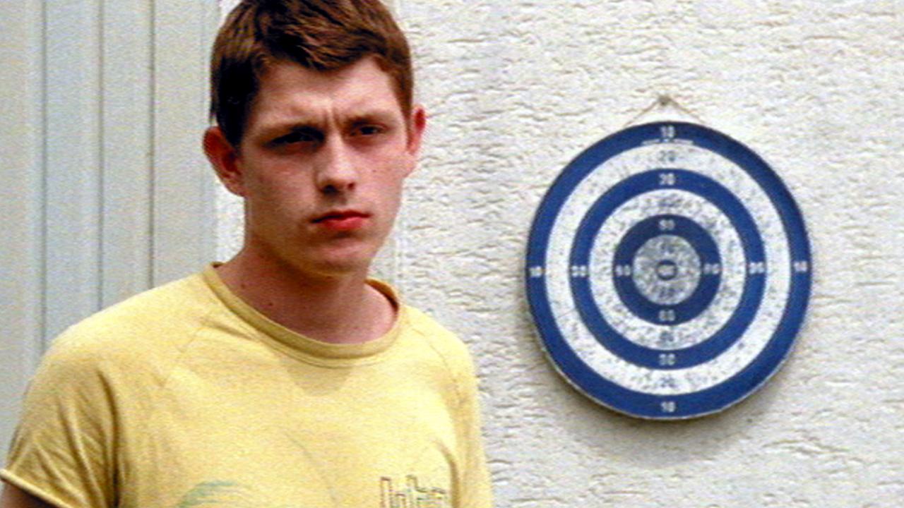 Szenenbild aus dem Drama BUGALOW: Ein junger Mann im gelben T-Shirt steht vor einer Hauswand. Rechts neben ihm hängt eine Darts-Scheibe.