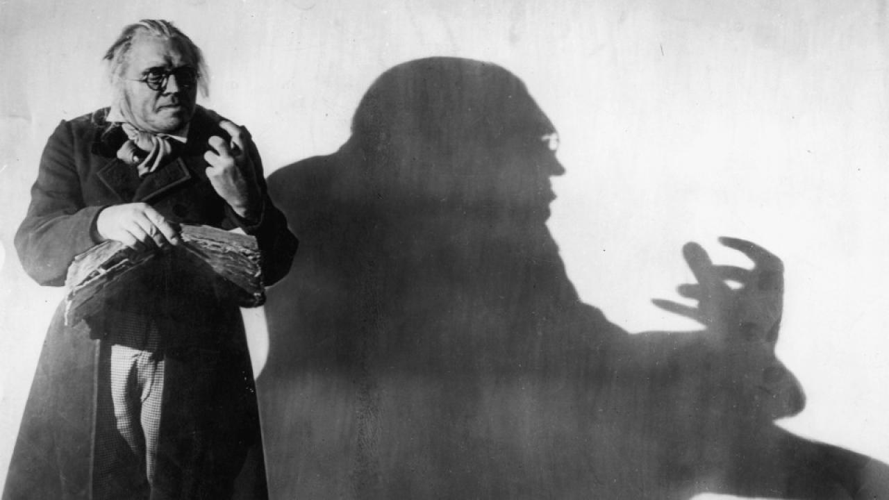 Das Cabinet des Dr. Caligari, Szenenbild: Mann in altmodischer Kleidung steht gegen einer weißen Wand. Sein Schatten ist groß und bedrohlich.