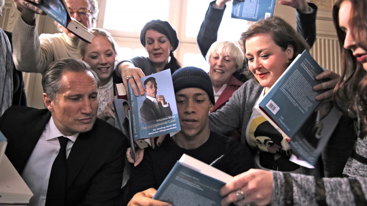 Heil, Szenenbild: Mehrere Menschen umringen einen Autor mit Büchern in der Hand, während er Autogramme in seine Werke schreibt.