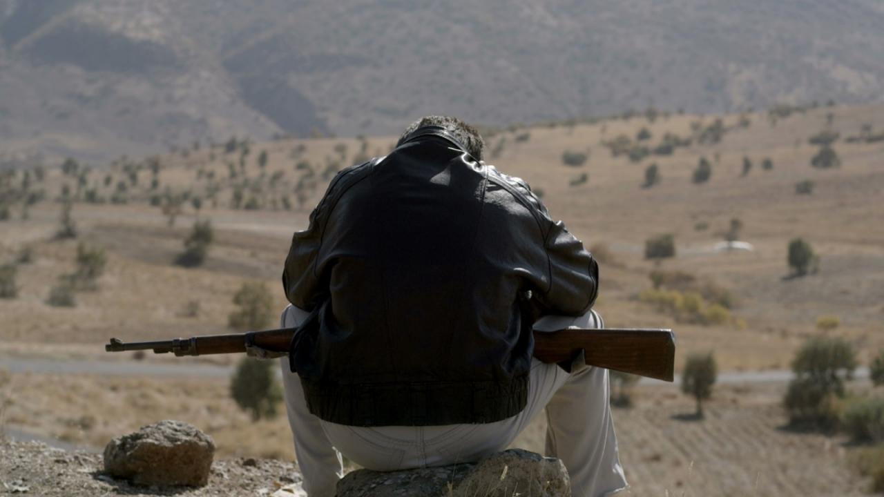 Der Junge Siyar, Szenenbild: Eine Figur von hinten sitzt auf einen Stein vor einer Wüstenlandschaft. Sie trägt eine schwarze Lederjacke und ein Gewehr liegt auf ihren Knien.