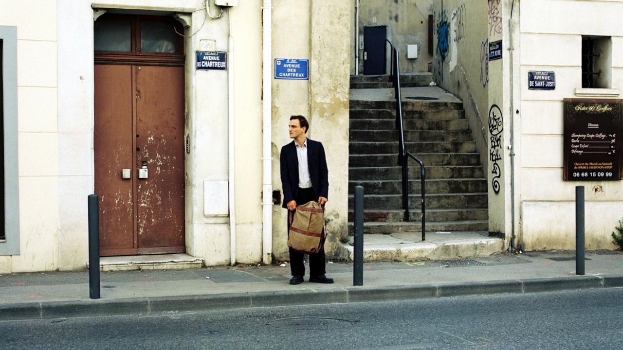 Szenenbild aus dem Drama TRANSIT: Georg, ein junger Mann, steht mit seinem Seesack vor einer Hauswand und einer Treppe in Marseille.