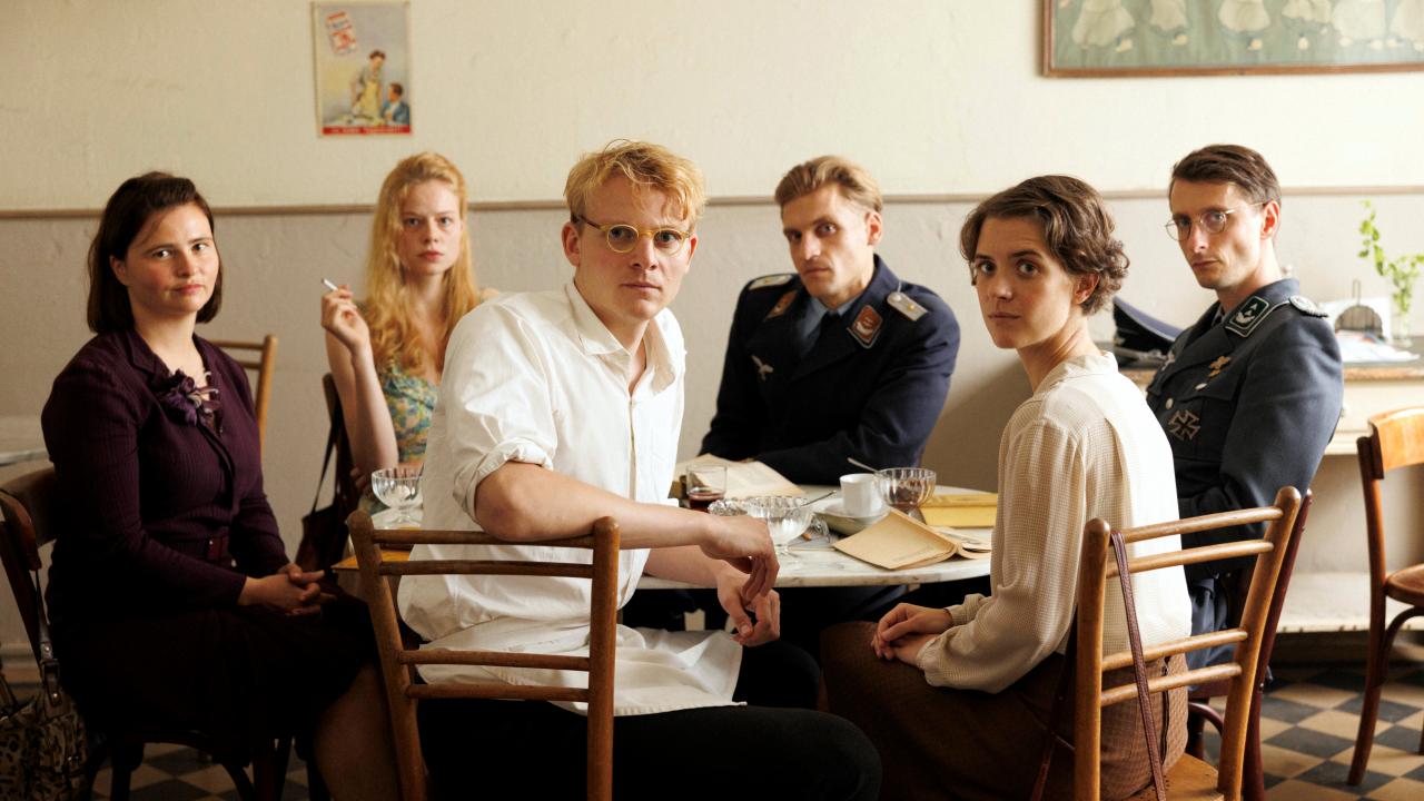 Szenenbild aus dem Drama IN LIEBE, EURE HILDE: Die jungen Widerstandskämpfer/-innen sitzen um einen Tischin einem Café, und blicken dabei in die Kamera. Im Vordergrund sieht man Hans und Hilde Coppi. Zwei der jungen Männer im Hintergrund tragen Uniform.