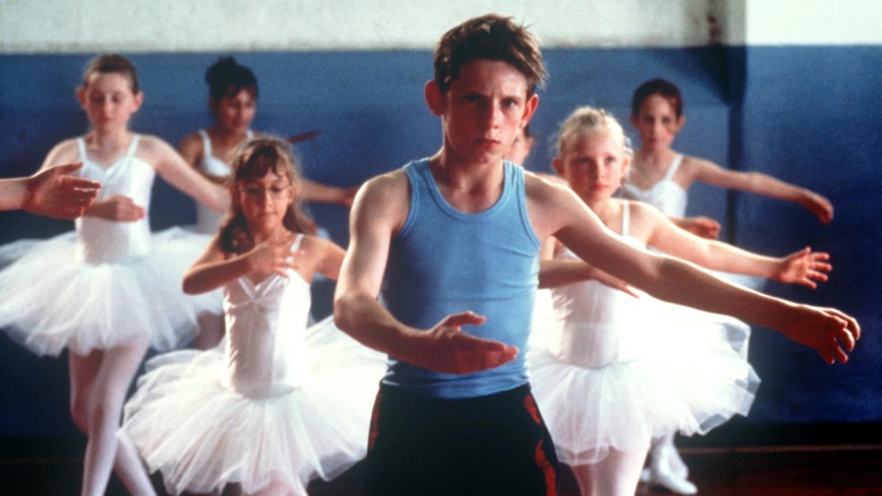 Szenenbild aus dem Film BILLY ELLIOT - I WILL DANCE: Ein Junge im blauen Unterhemd und Sporthose übt eine Balletposition. Hinter ihm fünf Mädchen in weißen Ballettkleidern.