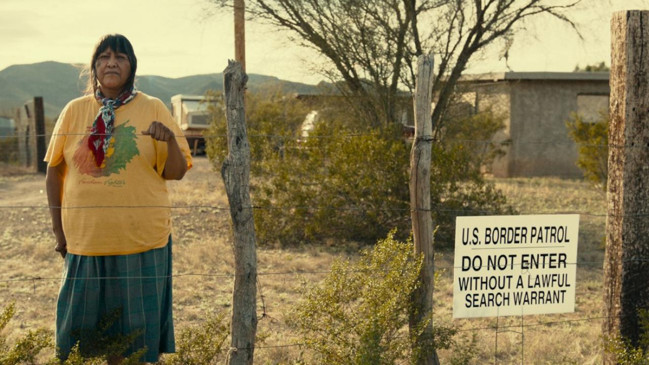 Borderland Blues, Szenenbild: Frau im gelben „Freedom Fighter“-Shirt steht hinter Stacheldrahtzaun neben einem Schild der U.S. Border Patrol in Wüstenlandschaft.