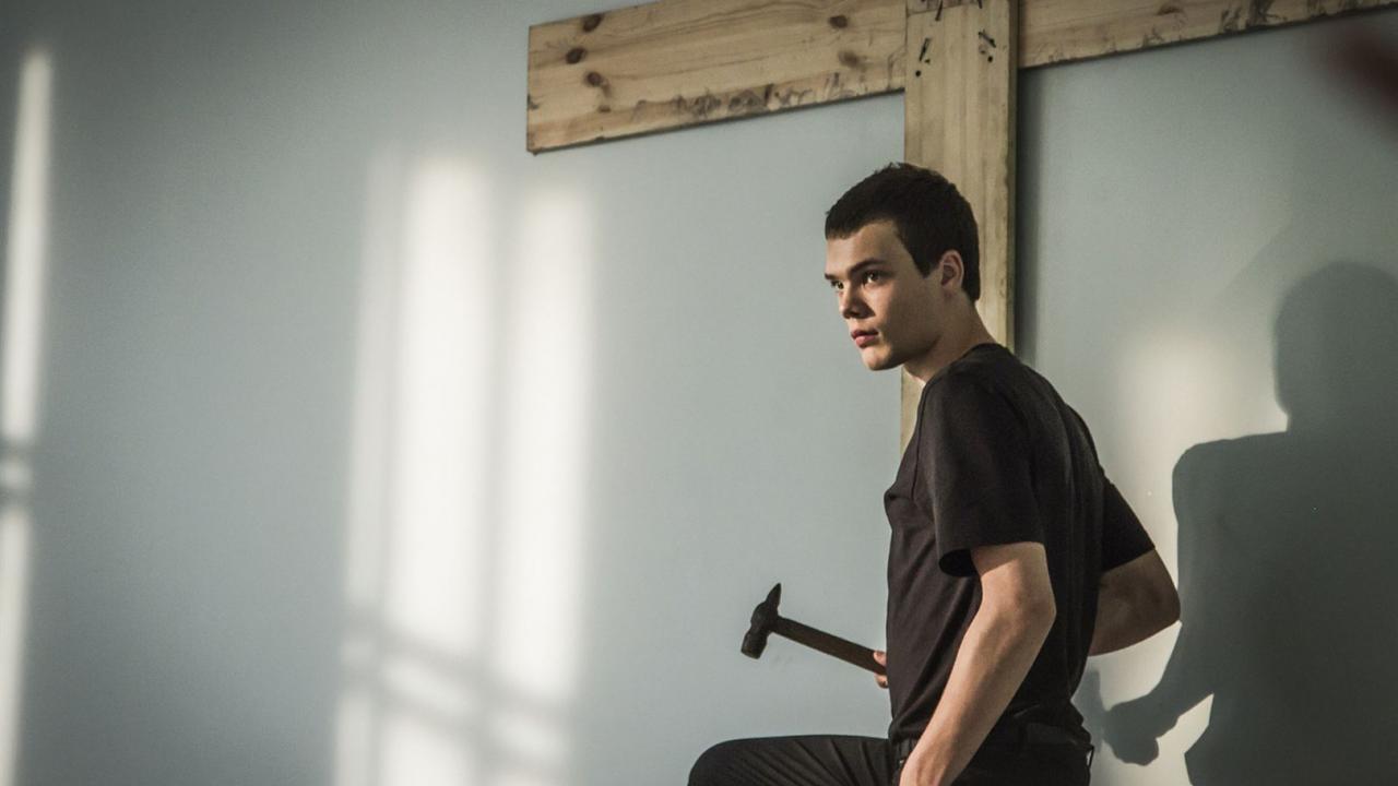 Der die Zeichen liest, Szenenbild: Junger Mann in schwarzem T-Shirt hält einen Hammer und steht vor einem großen Holzkreuz an der Wand.