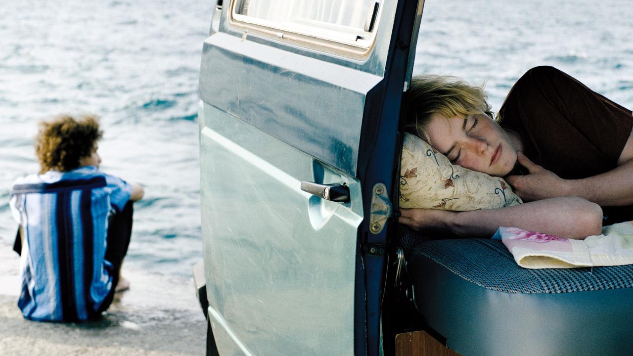 Szenenbild aus dem Film RÄUBERHÄNDE: Rechts im Vordergrund sieht man einen jungen Mann schlafen in einem Bus liegen. Im Hintergrund sitzt in der Rückenansicht ein anderer junger Mann am Wasser.