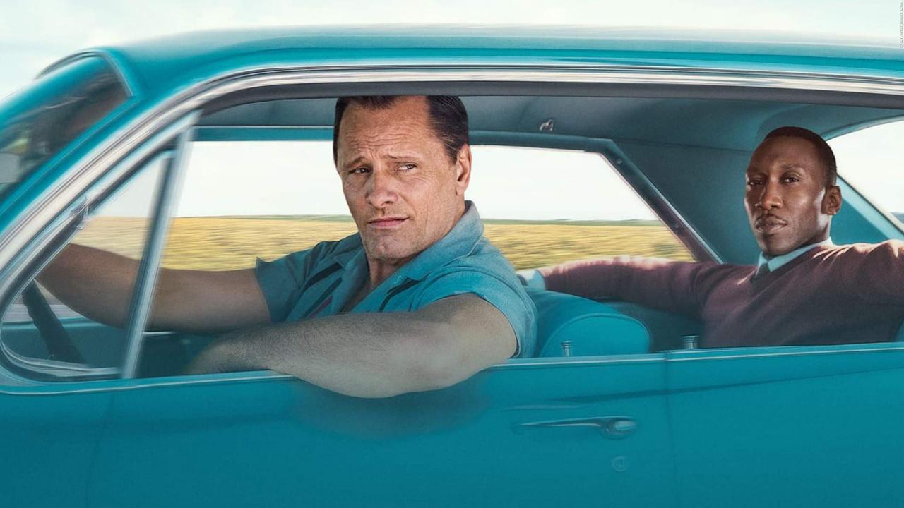 Szenenbild aus dem Drama GREEN BOOK: Halbtotale: Eine türkisfarbene Limosine aus den 50-/60er-Jahren. Der weiße Fahrer schaut aus dem Seitenfenster. Auf der Rückbank sitzt sein Schwarzer Fahrgast.