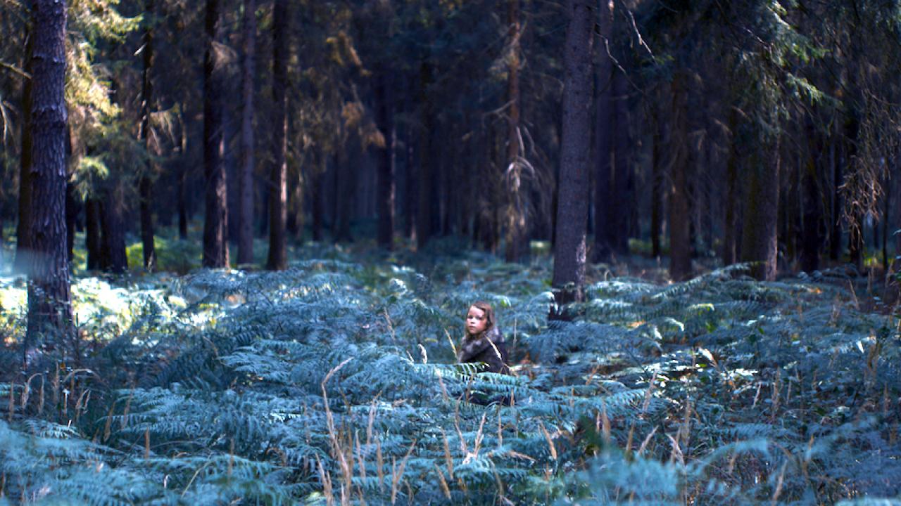 Szenenbild aus dem Drama MILCHZÄHNE: In einem Wald inmitten von Farnen steht ein kleines Mädchen und schaut in die Kamera.