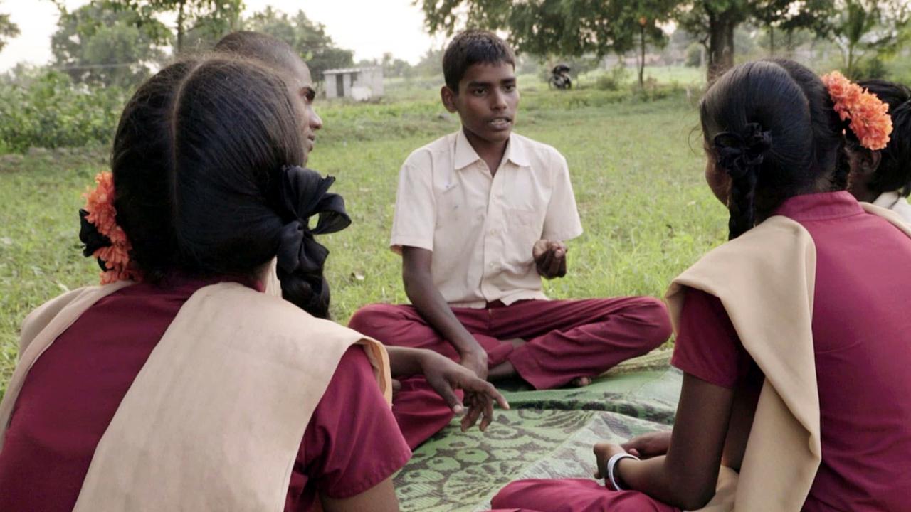 Szenenbild aus dem Dokumentarfilm POWER TO THE CHRILDREN: Drei Jungen und zwei Mädchen sitzen auf dem Rasen und diskutiere-