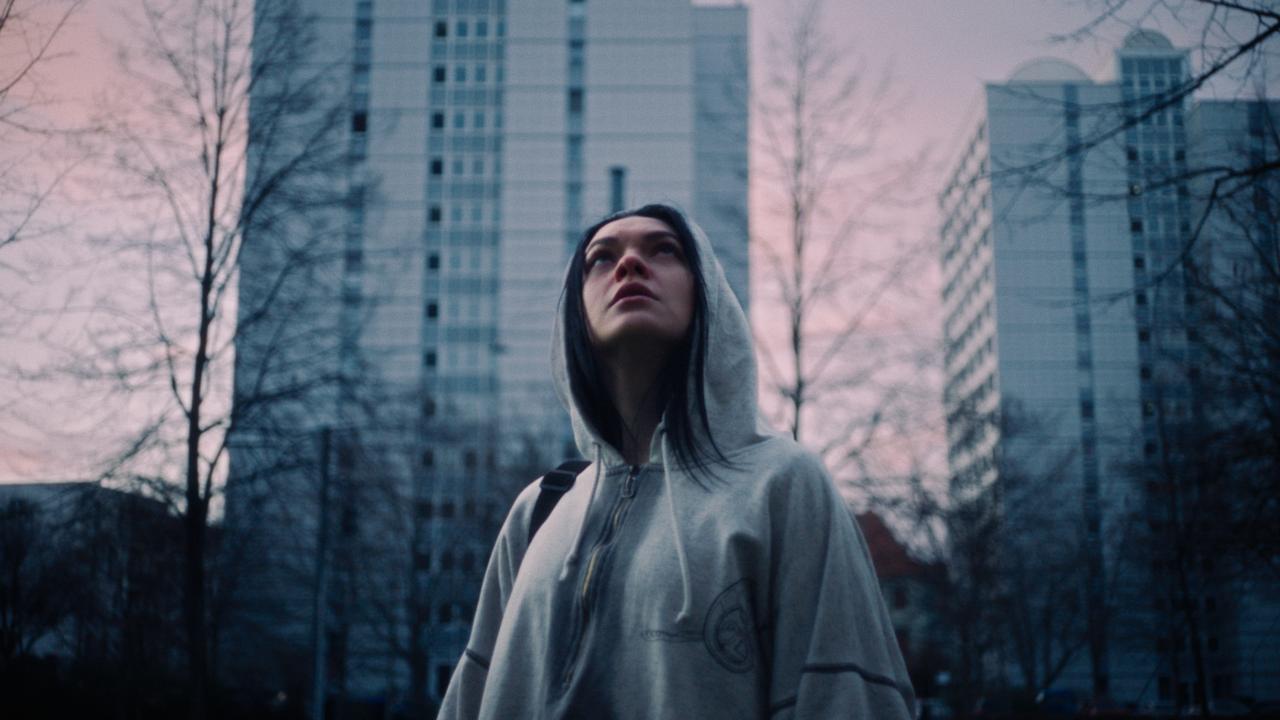 Szenenbild aus dem Drama VENA: Eine junge Frau im weißen Hoodie steht vor Hochäusern und schaut nach oben. Die Bäume sind kahl. Das Bild ist in grau-blauen Farben gehalten, der Himmel ist blass rosa.