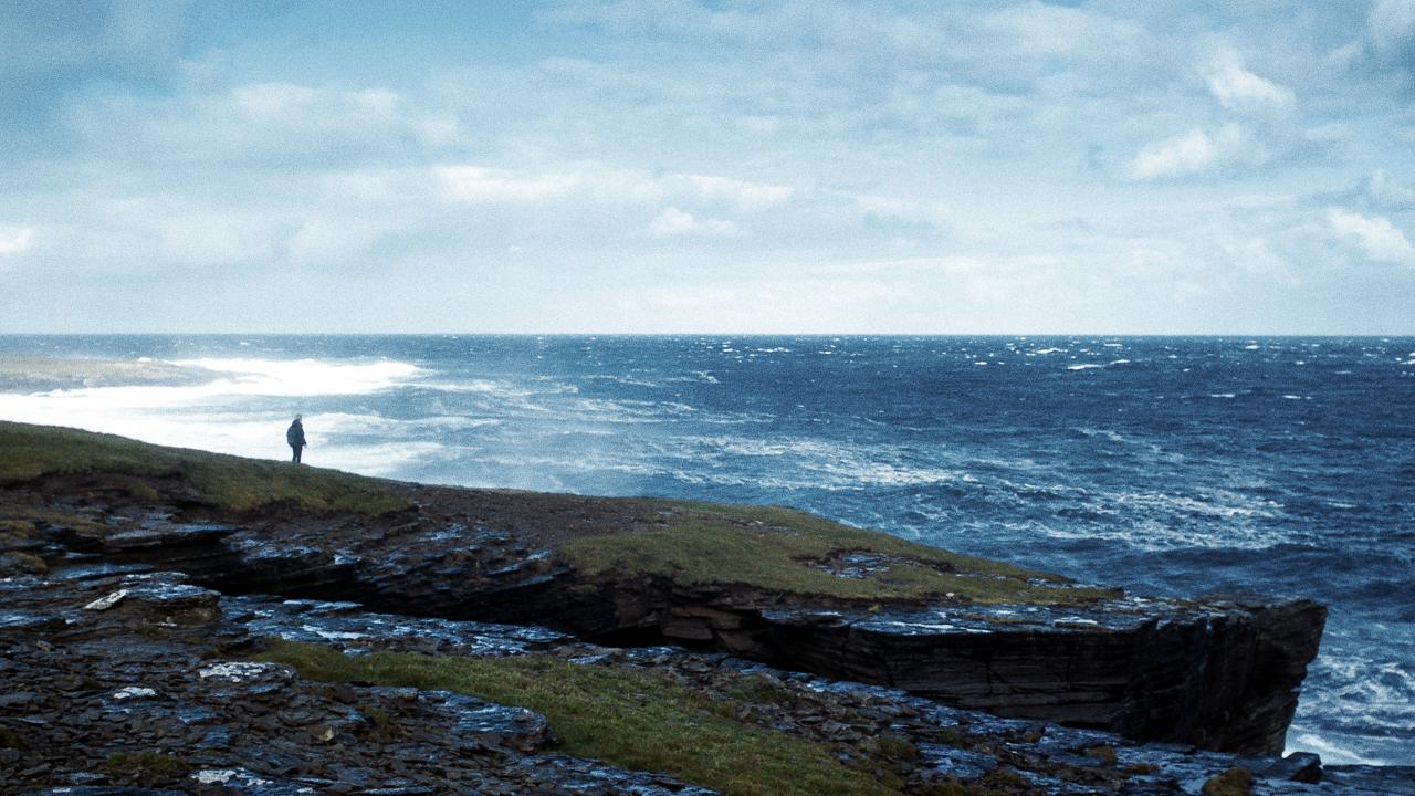 Szenenbild aus dem Drama THE OUTRUN: Panoramaaufnahme einer wilden Küstenlandschaft mit Blick auf. das Meer. Eine Figur steht klein am Rand der Klippen.