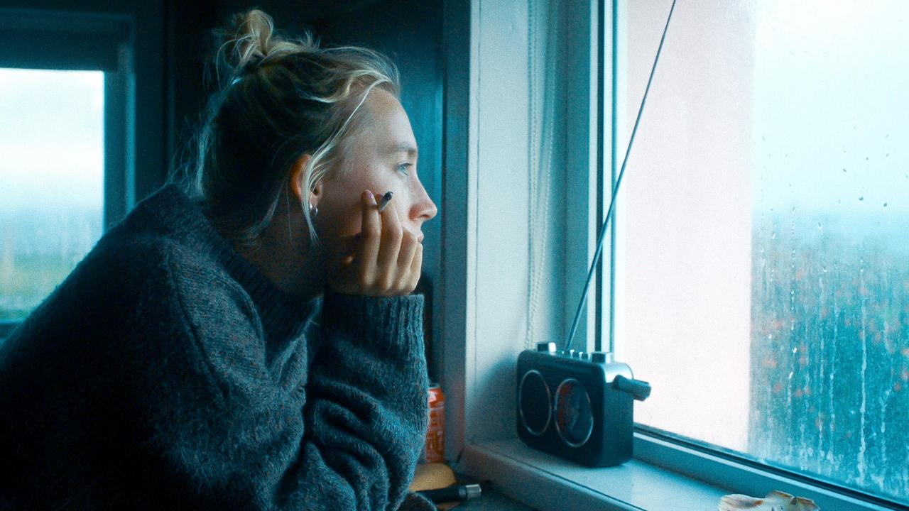 Szenenbild aus dem Drama THE OUTRUN: Porträt einer jungen Frau, die an einem Fenster steht und nach draußen in eine verregnete Landschaft schaut. Sie hält eine Zugarette in der Hand.Das Bild ist in blau-grauen Farbtönen gehalten.