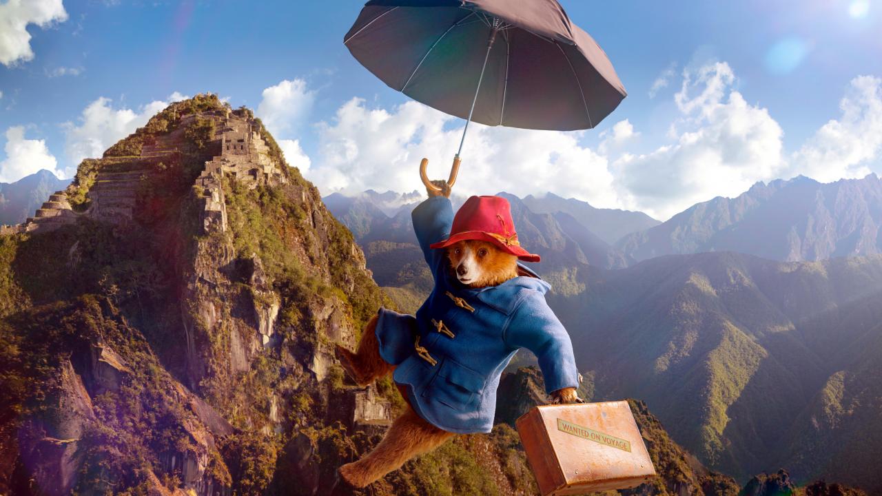 Filmstill aus dem Film PADDINGTON IN PERU: Der Bär Paddington hält sich an einem Regenschwirm und schwebt vor einer Berglandschaft im Himmel. Er trägt einen roten Hut, einen blauen Mantel und einen Koffer.