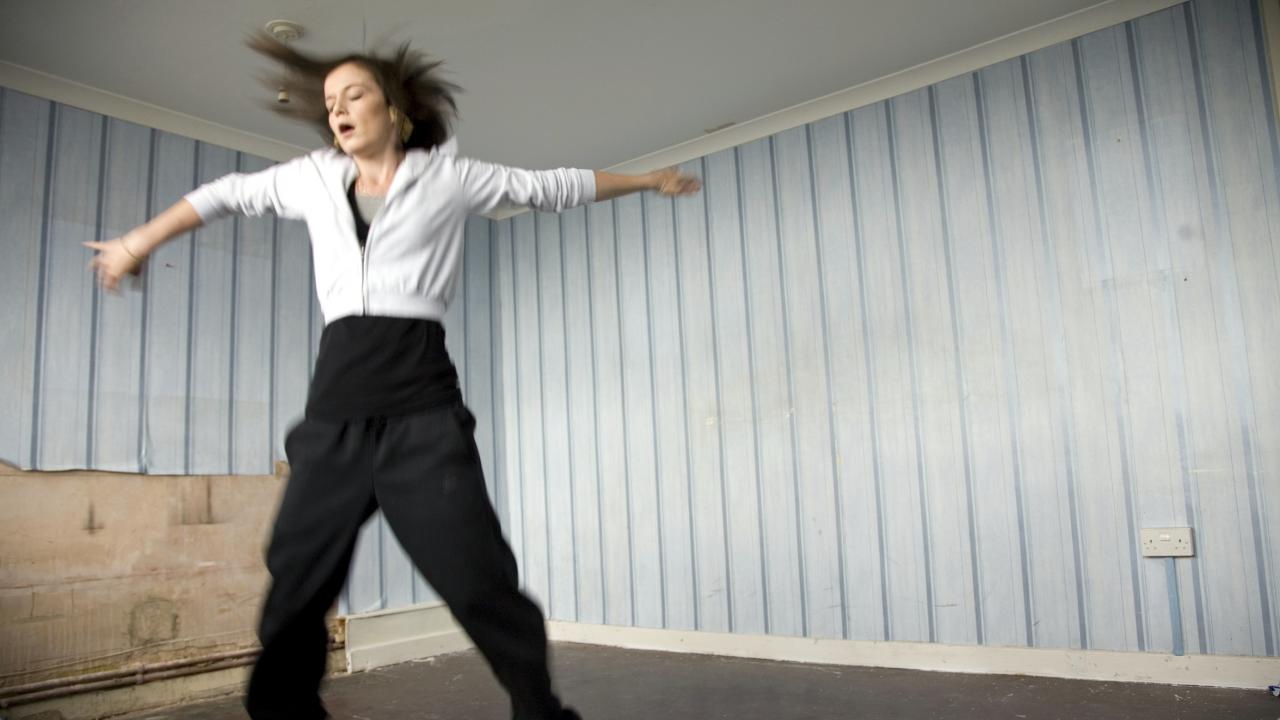 Filmstill aus dem Drama FISH TANK: Ein Mädchen im Teenageralter tanzt mit ausgestreckten Armen in einem kargen Raum. Sie trägt eine schwarze Jogginghose und eine weißes Oberteil. 