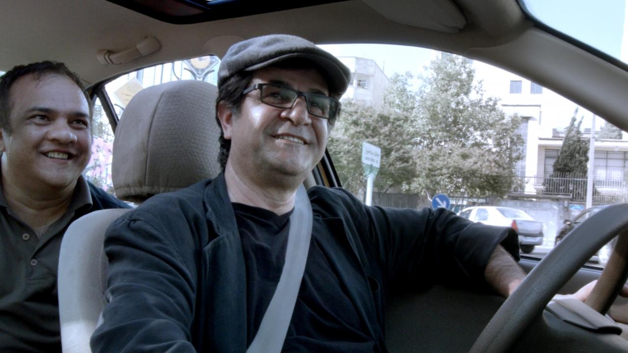 Szenenbild aus dem Dokumentarfilm TAXI TEHERAN: Der iranische Regisseur Jafar Panahi sitzt am lenkrad eines Autos und blickt lächelnd in die Kamera. Hinter ihm sitzt ein ebenfalls lächelnder Mann.