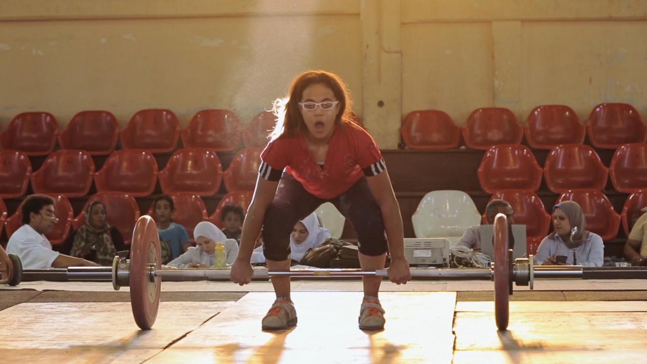 Lift Like a Girl, Szenenbild: Eine junge Sportlerin versucht in einer Turnhalle vor Publikum ein Gewicht zu stemmen.